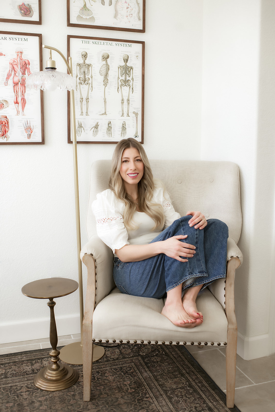 Brooke curled up barefoot in a cream armchair, anatomical charts and a brass lamp behind her